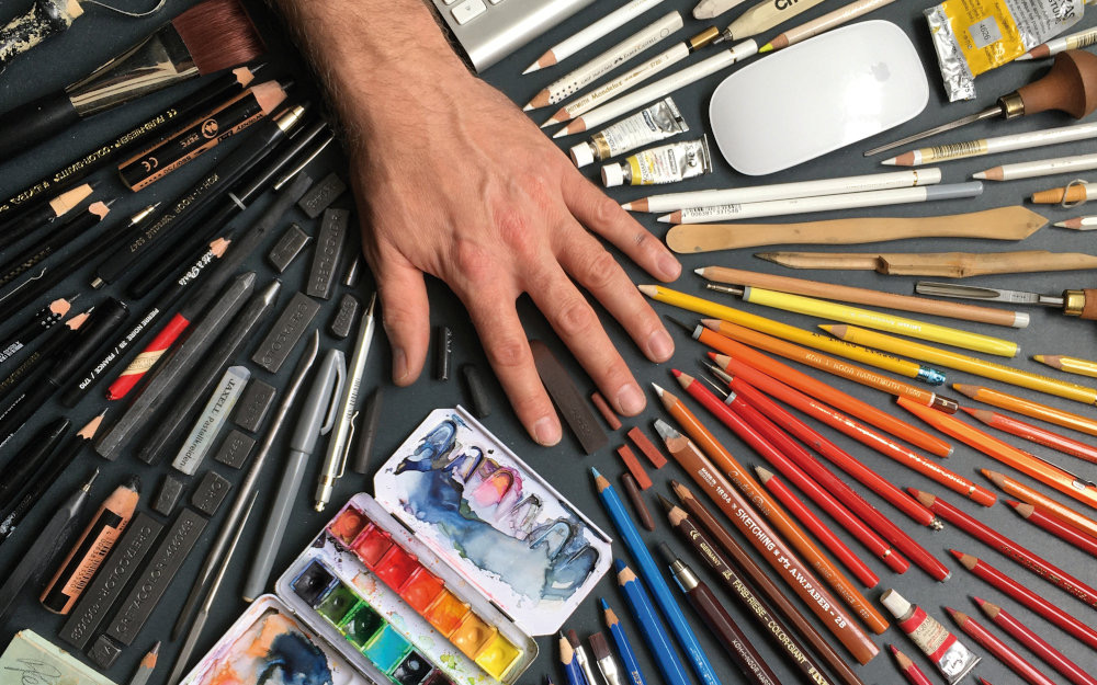 Zeichenmaterial auf dem Tisch: Stifte, Kreiden, Pinsel und ein Aquarellkasten, darauf eine Hand